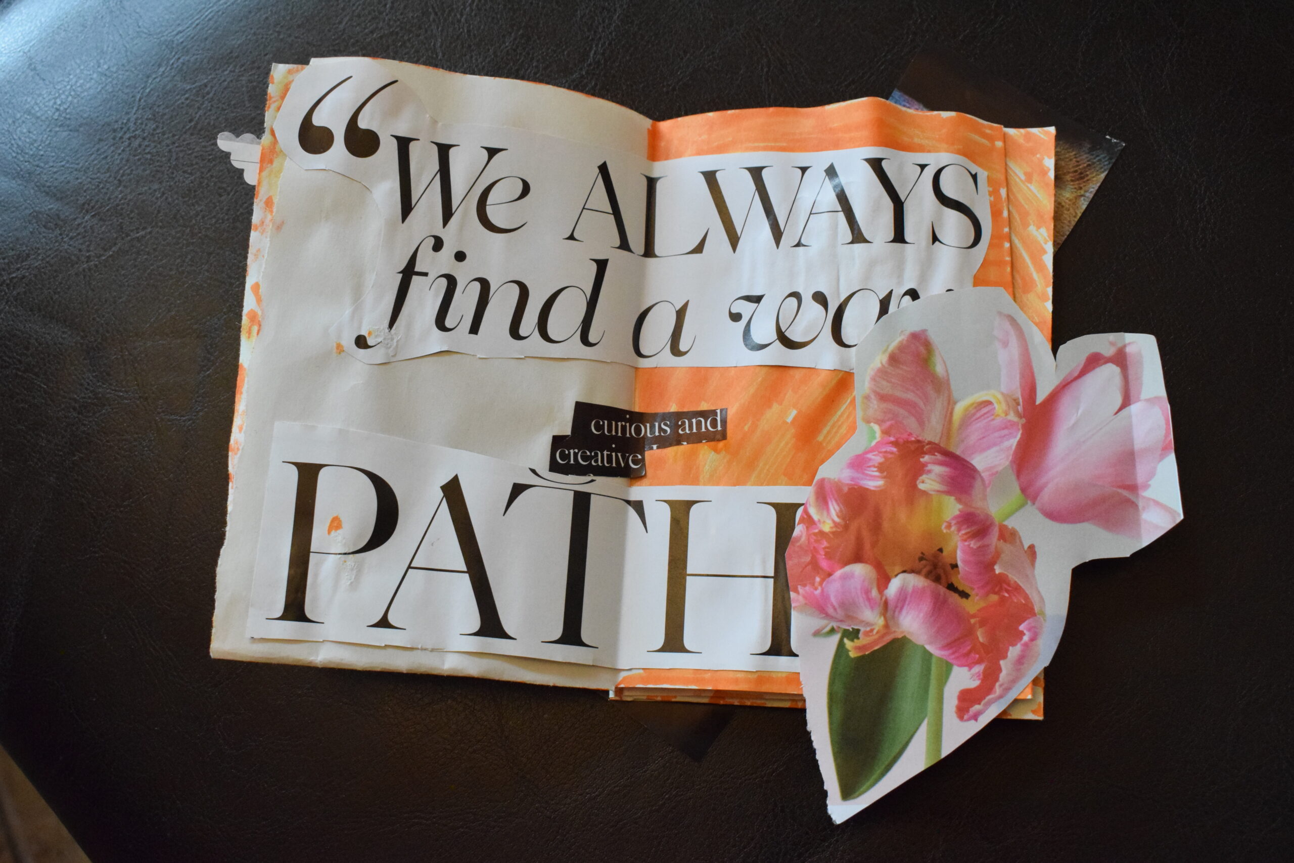 a zine sits on a dark leather surface with cut our magazine letters that read 'we always find a way, curious and creative path' with a cut out of flowers over top.