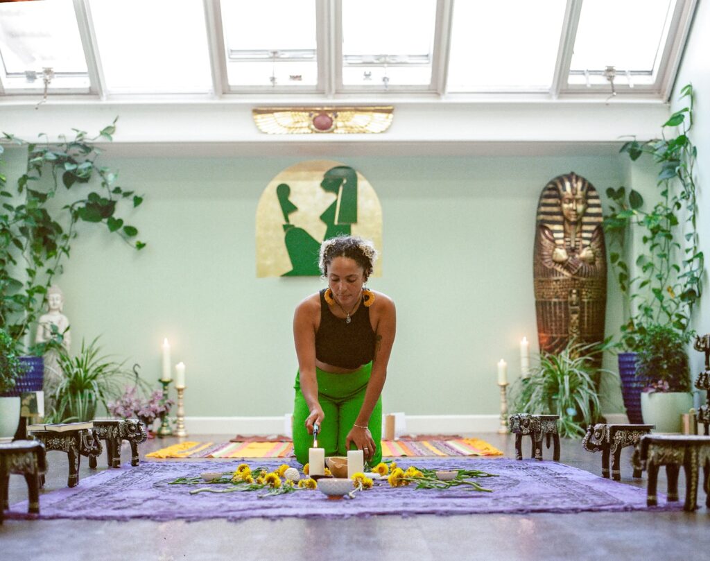 Image of Bekah kneeling on mat holding a ceremonial space. Surrounded by plants.