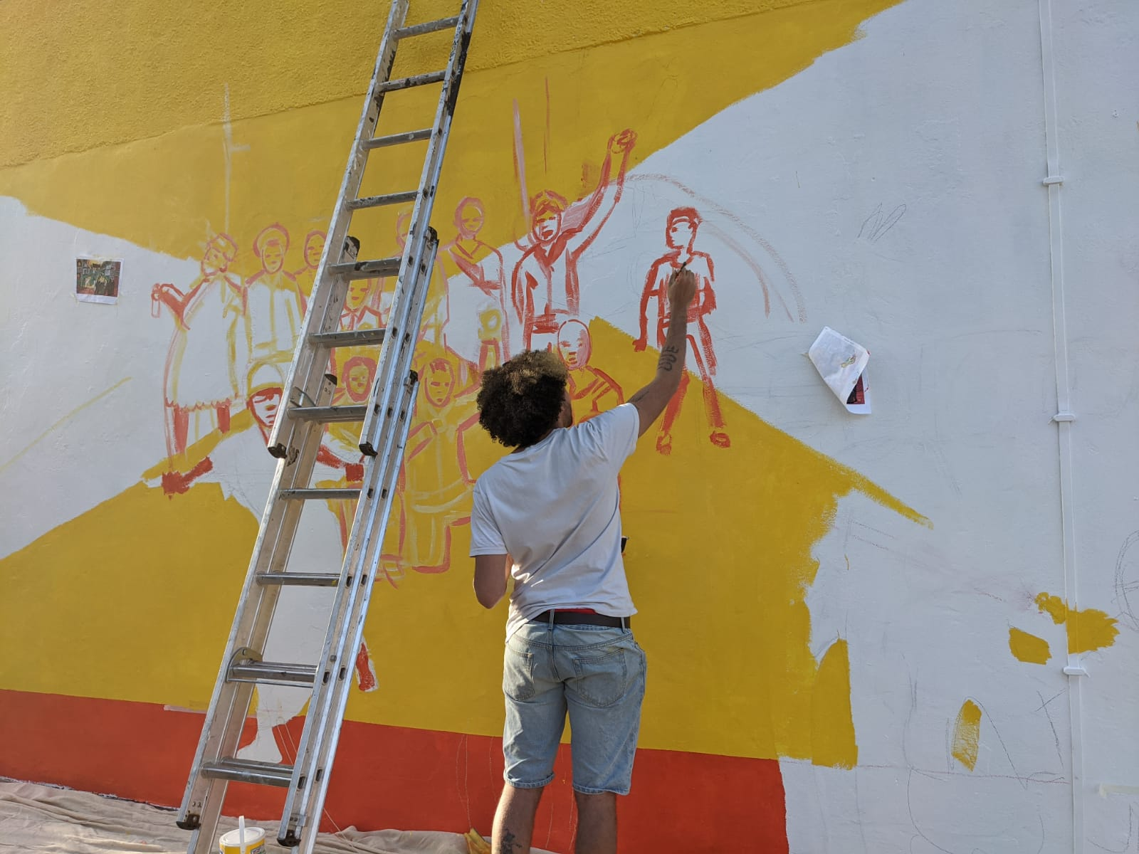 Jacob V Joyce stands with his back to the camera painting an unfinished yellow and red mural. A ladder stands beside them