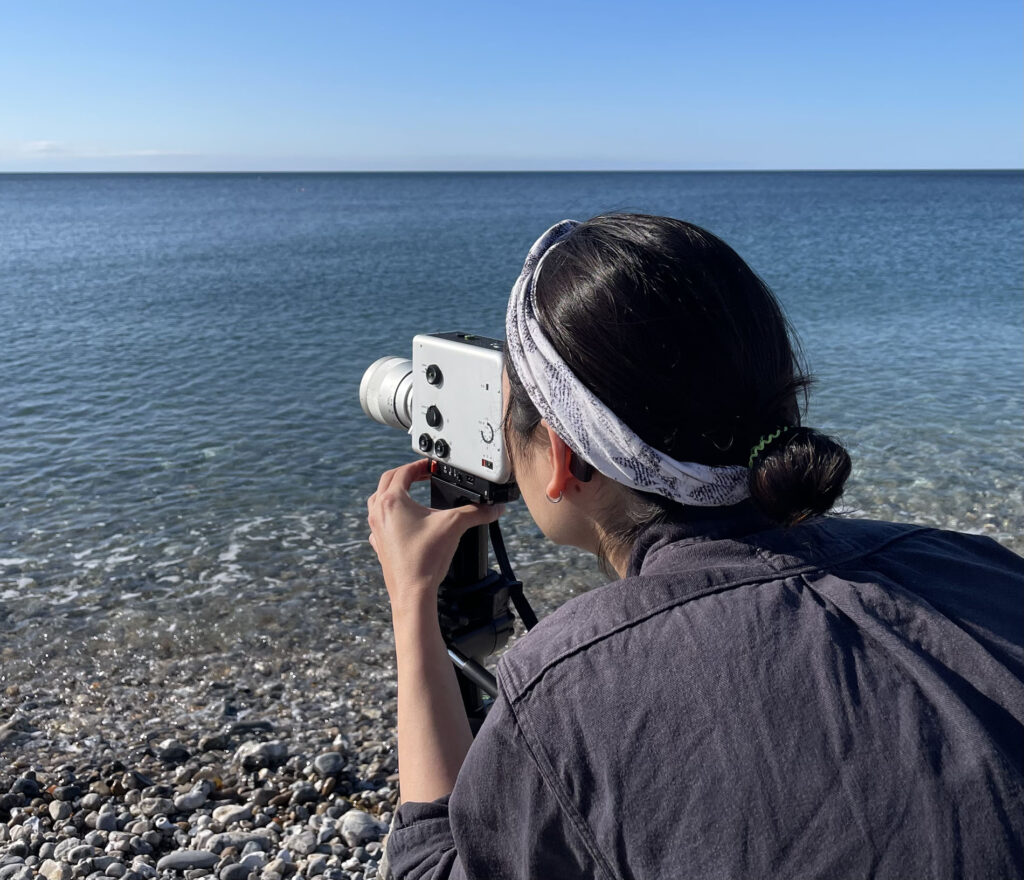 A photo of Seo Hye with her back to the camera, facing a ocean horizon with a pebbly grey beach. She has her black hair tied back into a bun and is wearing a grey and speckled black headband. She is holding a analogue camera to her face as she photographs the sea.