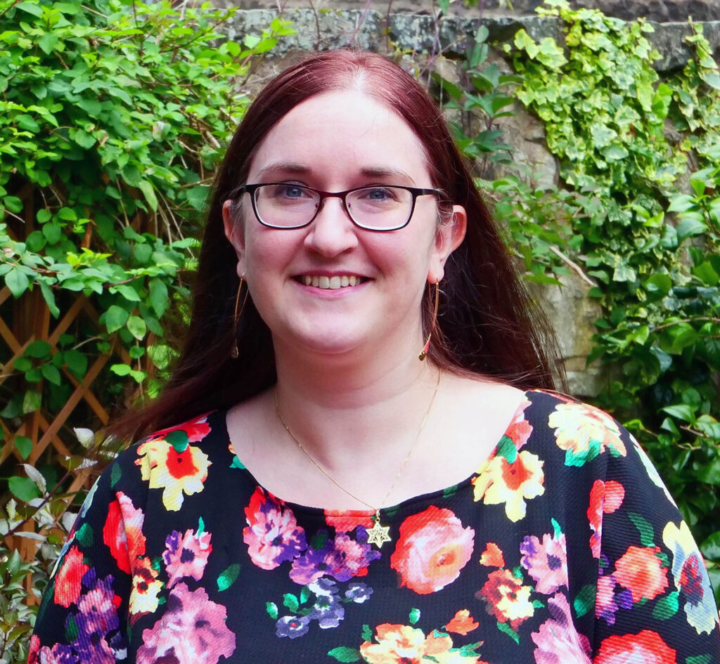 A photo of Tzipporah facing the camera, with a smile on her face. Tzipporah has long brown hair, black thin framed glasses, long dangly earrings and wearing a pink, purple and yellow floral top. She is standing against a garden wall with green plants and foliage in view