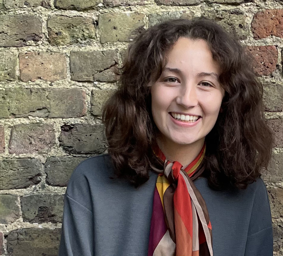 A photo of Kaiya facing the camera with her back against a brick wall. She has medium length brown wavey hair, a big smile on her face and is wearing a grey jumper with a red, yellow, purple and grey neckerchief.