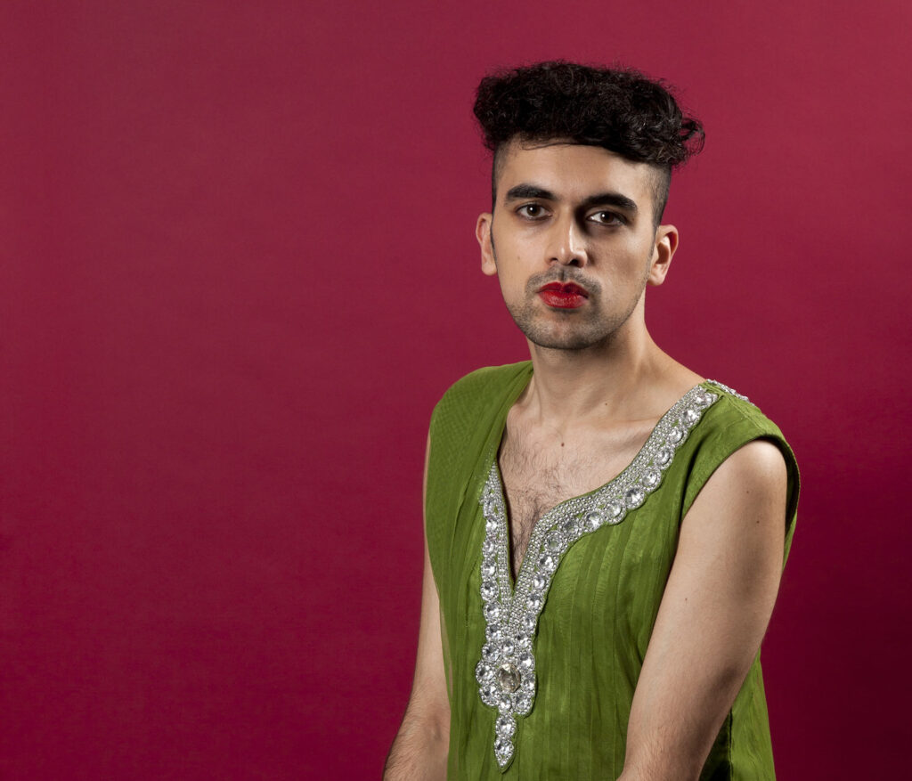 This is a photo of Vijay taken by Holly Revell. Vijay is a brown person with black hair. He is wearing a leafy green short sleeve saree with jewell detailing. He has red lipstick on and black eyeliner. The background is red. He is sitting down and his hands are placed together on his lap.