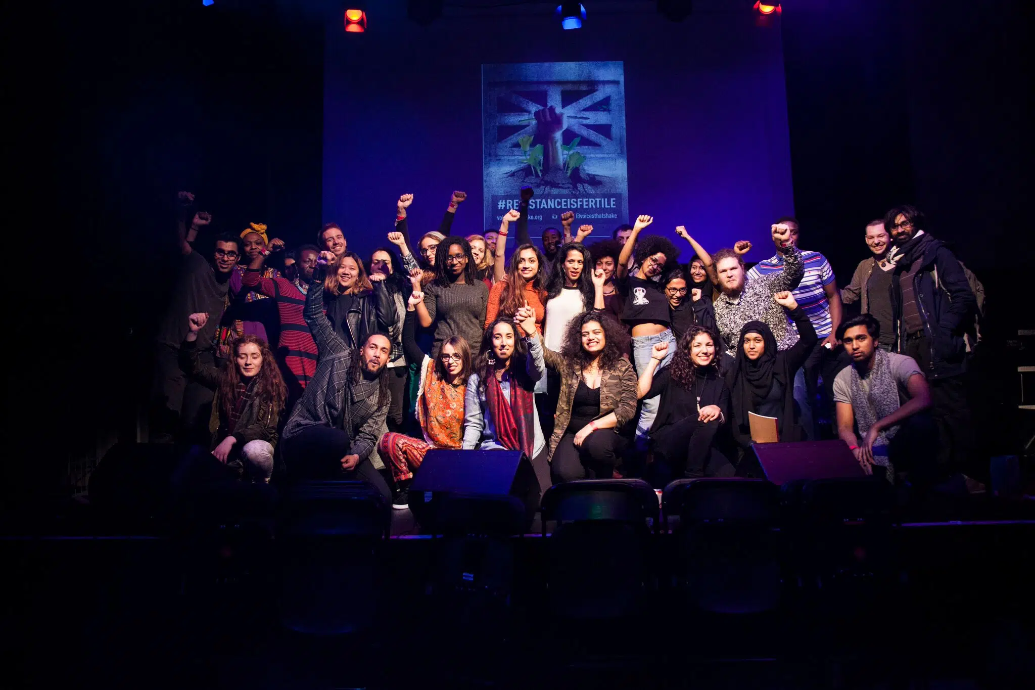a group of people stand and sit on a stage. many of them have a raised fist